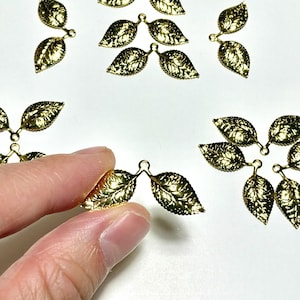May include: A collection of small, gold-colored metal leaf charms, each featuring two connected leaves with detailed vein textures. The charms are arranged in various groupings, with one individual piece held up to show the design clearly.