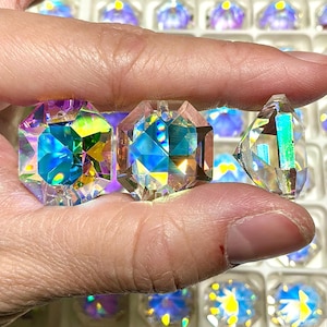 Puede incluir: Primer plano de tres pedrerías de cristal facetadas e iridiscentes sostenidas en una mano. Los cristales tienen forma geométrica y reflejan un espectro de colores, incluyendo azul, verde y amarillo. El fondo muestra una bandeja llena de cristales similares.