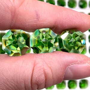 Puede incluir: Tres gemas verdes octogonales facetadas se sostienen entre los dedos. Las gemas tienen un agujero perforado en la parte superior. El fondo está lleno de más gemas verdes en un patrón de cuadrícula.
