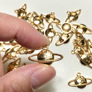 May include: Gold-colored planet charms with a star on the planet's surface and a ring around it. The charms are small, with a loop at the top for attaching to jewelry or other crafts. The charms are scattered on a white surface.