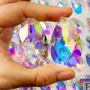 May include: Two iridescent crystal teardrop pendants with faceted edges. The pendants are held in a hand against a background of many similar pendants.