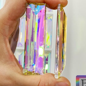 May include: Two iridescent crystal prisms, one is longer than the other, both have a rectangular shape with beveled edges. The prisms are held in a hand.