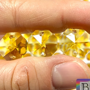 May include: Three amber-colored crystal octagon beads with two holes each. The beads are held in a hand against a background of many similar beads.