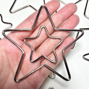 May include: A collection of silver-colored star-shaped wire forms. The stars vary in size, with one nested inside another. The wire is smooth and reflective, creating a bright, metallic appearance. The stars are arranged on a white surface.