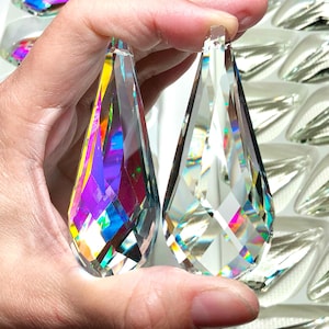May include: Two clear, faceted crystal teardrop pendants are held up, showcasing their intricate cuts and rainbow iridescence. Additional crystal pendants are visible in the background, arranged in a tray. These decorative crystals are ideal for chandeliers or other craft projects.