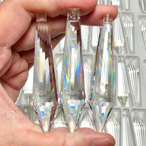 May include: Three clear crystal teardrop prisms of varying sizes, held in a hand. The prisms are faceted and reflect light, creating a rainbow effect.