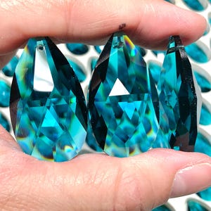 Puede incluir: Una mano sostiene tres colgantes de cristal facetados, en forma de lágrima, de color verde azulado. Los cristales tienen múltiples facetas que reflejan la luz, creando un efecto brillante. Cristales verde azulado adicionales son visibles en el fondo.