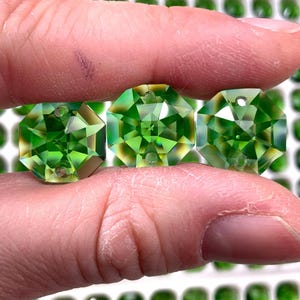 May include: Three faceted, octagonal green gemstones are held between fingers. The gems have a bright, reflective quality, with multiple facets catching the light. The gems are a vibrant, clear green color, and appear to be of high quality.