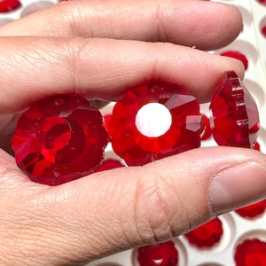 May include: Close-up of several faceted, ruby red crystal flower-shaped beads. Each bead has a white centre and a hole for stringing. The beads are held in a hand, with many more visible in a white tray.