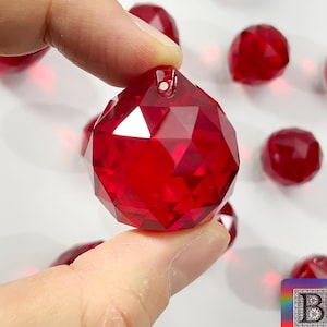 Puede incluir: Primer plano de un adorno de bola de cristal roja con un agujero para colgar. El adorno es facetado y tiene un acabado brillante.