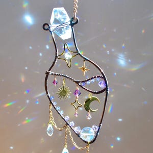 May include: A handcrafted suncatcher in the shape of a bottle, made with copper wire and adorned with celestial charms. Features a large crystal at the top, dangling crystals, and gold-colored stars and a crescent moon. The suncatcher is designed to catch and refract light.