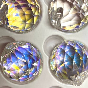 May include: A close-up of six iridescent crystal balls in a white foam packaging. The balls are faceted and reflect light in a rainbow of colors.