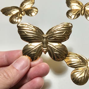 May include: Several gold-colored butterfly embellishments are displayed on a white surface. The butterflies have detailed wing patterns and are in various positions. One butterfly is being held by a hand.