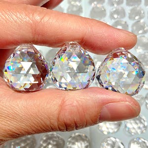 May include: Three clear crystal prism pendants with faceted surfaces are held between fingers. The prisms are teardrop-shaped and reflect a rainbow of colors. Additional prisms are visible in the background.