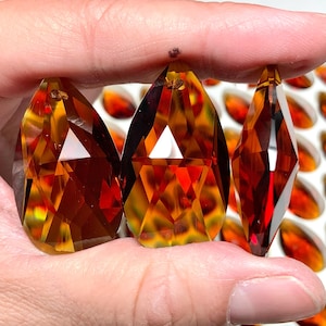 Könnte beinhalten: Drei facettierte, bernsteinfarbene Kristallanhänger in Tropfenform werden gezeigt. Die Kristalle haben ein geometrisches Design mit einem Loch oben zum Aufhängen. Der Hintergrund zeigt weitere bernsteinfarbene Kristalle.