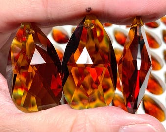 May include: Three faceted amber-colored crystal pendants with a teardrop shape are held up. The crystals have a geometric design with a hole at the top for hanging. The background shows more amber-colored crystals.