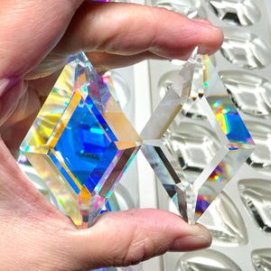 Può includere: Due pendenti di cristallo trasparente sfaccettati a forma di diamante sono tenuti in mano, mostrando una vibrante esposizione di colori arcobaleno. Ulteriori forme di cristallo sono visibili sullo sfondo, suggerendo una collezione di elementi decorativi.