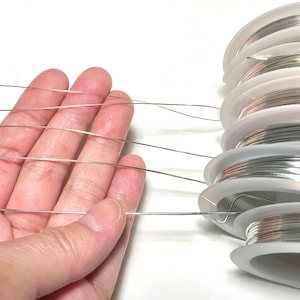 Peut inclure: Gros plan d'une main tenant plusieurs bobines de fil d'argent. Le fil est fin et semble être destiné à la fabrication de bijoux.