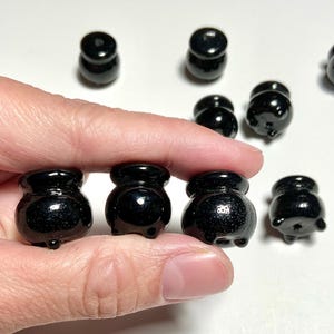 May include: A collection of small, glossy black cauldron-shaped figurines. Some are held in a hand, while others are scattered in the background. Each cauldron has a rounded body, a flared rim, and three small feet. The figurines are made of a reflective material.