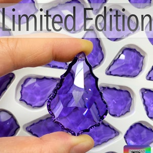 Puede incluir: Un colgante de cristal púrpura facetado en forma de lágrima se sostiene entre los dedos, con otros cristales similares en el fondo. Las palabras "Limited Edition" están en la parte superior. El logotipo de Bitsy Supply está en la esquina inferior derecha.