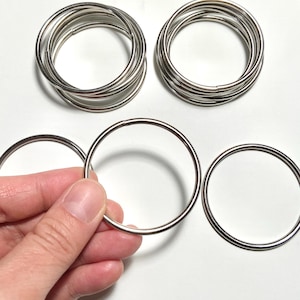 May include: Several stacks of silver metal rings, with one ring held in a hand. The rings are round and appear to be made of a shiny metal. The background is white.