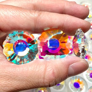 May include: Close-up of iridescent crystal flower-shaped beads with a central blue hue. The beads reflect a spectrum of colours, including orange, pink, and teal. A clear, faceted crystal bead is also visible. These beads are likely for jewellery making or craft projects.