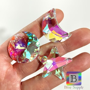 May include: Three iridescent crystal shapes: a circle, a crescent moon, and a five-pointed star. The crystals reflect a rainbow of colors, including pink, yellow, and blue. The shapes are held in a hand.