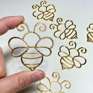 May include: A collection of gold-coloured bee-shaped charms. The charms are crafted from thin wire, featuring intricate designs such as antennae, wings, and striped abdomens. One charm is held by a hand, with others scattered on a white surface.