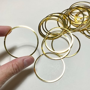 May include: A collection of gold-colored metal rings, some stacked together. One ring is held by a hand. The rings are thin and circular, with a shiny finish. The rings are likely for jewelry making or craft projects.