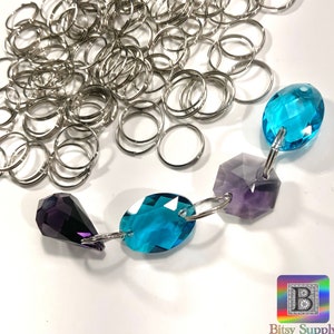 Puede incluir: Primer plano de una cadena de cuentas de cristal, que incluye un cristal morado en forma de lágrima, un cristal azul verdoso redondo, un cristal de amatista hexagonal y un cristal azul verdoso redondo. Los cristales están conectados por anillos de salto plateados. Hay una pila de anillos de salto plateados en el fondo.