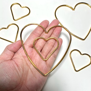 May include: A collection of gold-coloured wire heart shapes in different sizes. One large heart shape contains a smaller heart. The hearts are arranged on a white surface. The hearts are made of a shiny, metallic material.