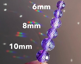 May include: A strand of faceted, purple beads in 6mm, 8mm, and 10mm sizes. The beads are strung on a thin wire and held by a person. The background has light reflections and the text "6mm", "8mm", and "10mm".