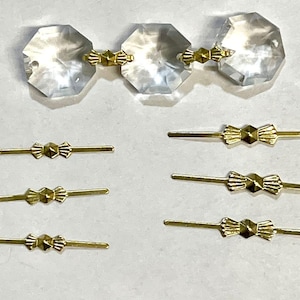 Może przedstawiać: Kolekcja złotych i srebrnych łączników w kształcie kokardki do tworzenia biżuterii. Łączniki są ułożone w rzędach na białym tle. W środku obrazu znajdują się trzy przezroczyste kryształowe koraliki.