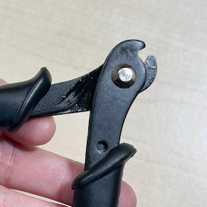 Op de afbeelding: Een zwarte draadkniptang met gebogen handgrepen en een metalen scharnierpunt. Het gereedschap heeft een matzwarte afwerking op de handgrepen en een kleine ronde opening aan het uiteinde van één handgreep. De snijbladen bevinden zich bovenaan.
