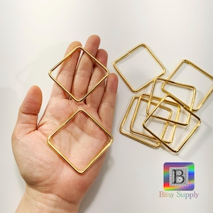 May include: A close-up of a hand holding several gold-colored metal square rings. The rings are all the same size and shape, and they are arranged in a pile.