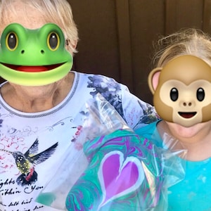 May include: Two people are holding a clear plastic bag with a green, purple, and white marbled heart-shaped object inside. The person on the left is wearing a white shirt with a hummingbird design and the person on the right is wearing a turquoise shirt.
