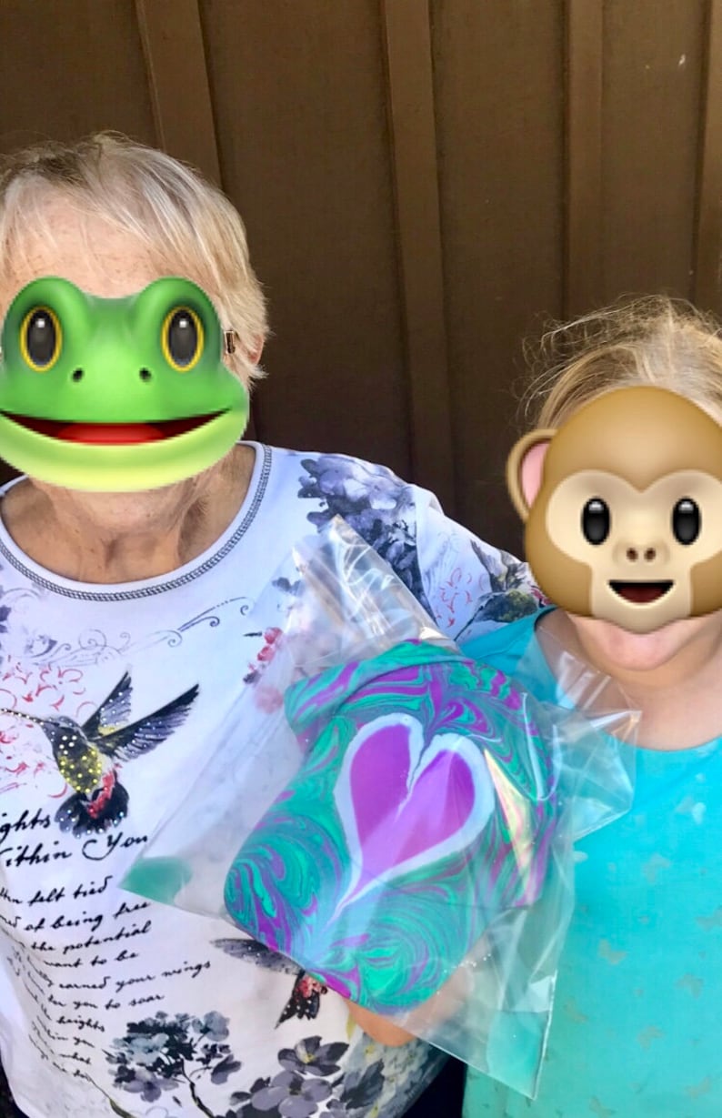 May include: Two people are holding a clear plastic bag with a green, purple, and white marbled heart-shaped object inside. The person on the left is wearing a white shirt with a hummingbird design and the person on the right is wearing a turquoise shirt.
