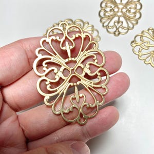 May include: Gold-colored filigree oval pendant held in a hand, with intricate openwork design. The pendant features a symmetrical pattern of swirling lines and floral elements. Several similar pendants are visible in the background.