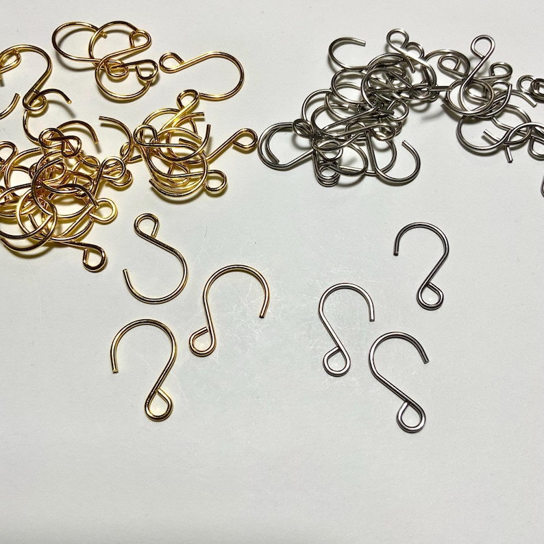 May include: An assortment of gold and silver-colored earring hooks and loops. The hooks are in various shapes, including S-shaped and curved designs. The loops are tightly wound and clustered together.