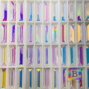 Peut inclure: Une feuille de strass rectangulaires en verre iridescent dans diff&eacute;rentes nuances de bleu, violet et rose. Les strass sont dispos&eacute;s en rang&eacute;es et en colonnes sur un fond blanc.