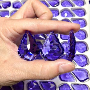 May include: Three faceted, teardrop-shaped, purple crystal pendants are held in a hand. The crystals have multiple facets that reflect light. The background shows more purple crystal pendants in a white tray. These pendants are suitable for jewelry making.
