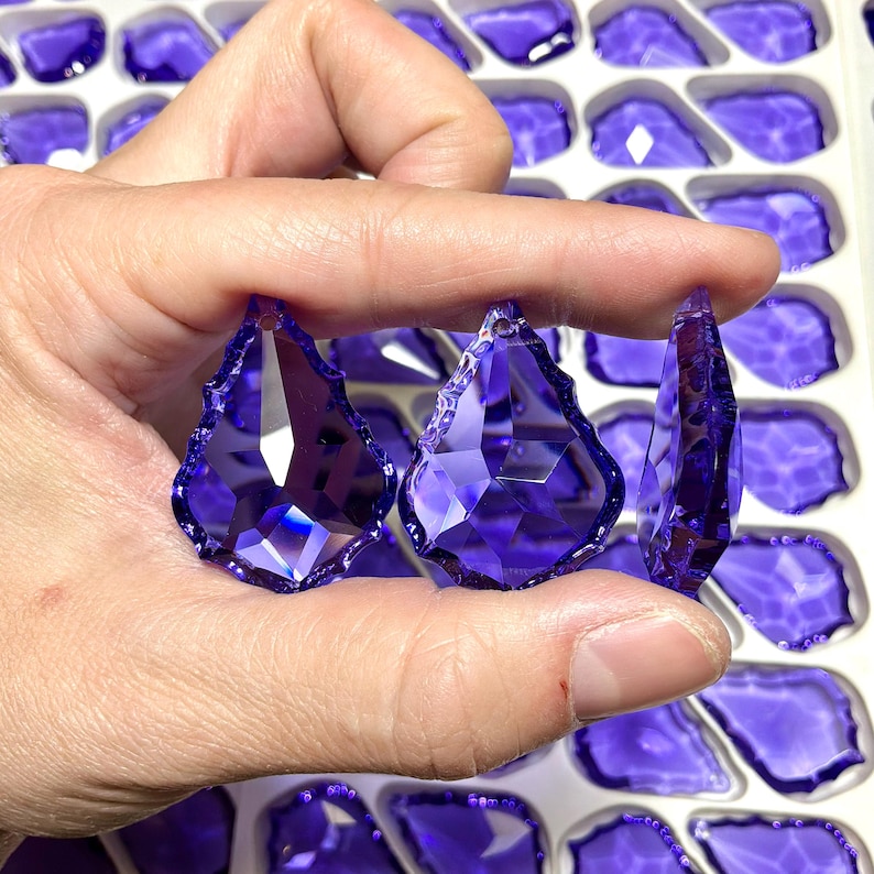 May include: Three faceted, teardrop-shaped, purple crystal pendants are held in a hand. The crystals have multiple facets that reflect light. The background shows more purple crystal pendants in a white tray. These pendants are suitable for jewelry making.