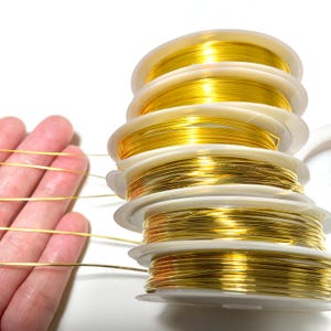 May include: A stack of spools of gold-colored wire, each spool is wrapped in white plastic. The wire is thin and appears to be for jewelry making.