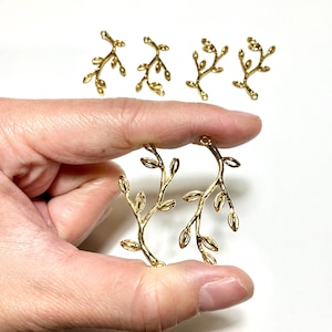 May include: Six gold-toned metal charms shaped like delicate tree branches with small leaves. Two of the branch charms are held in the foreground, while four identical branch charms are arranged in a row in the background.
