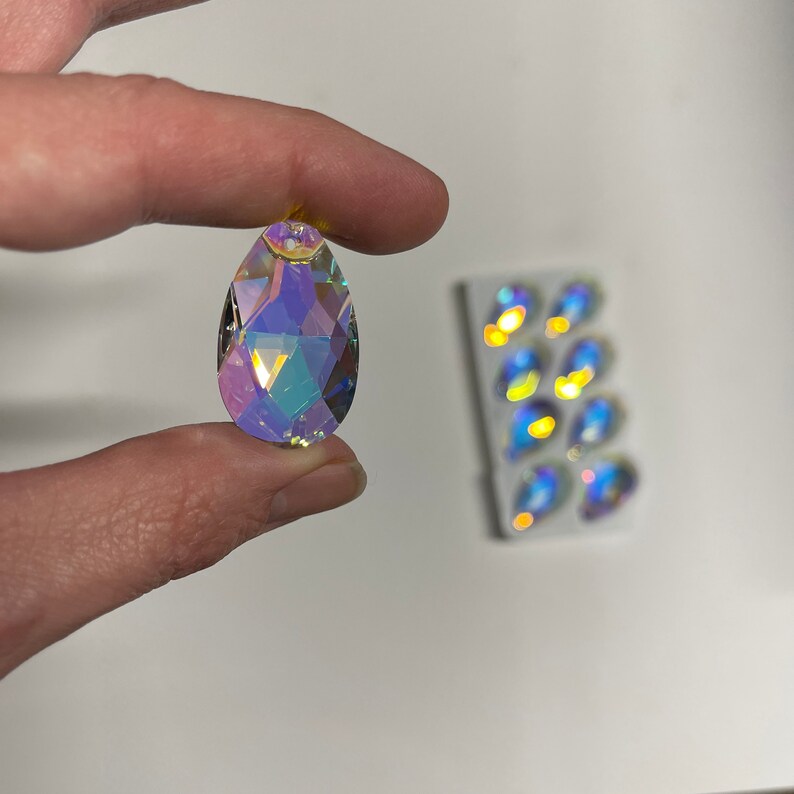 May include: A teardrop-shaped iridescent crystal prism with a faceted design. The prism is held in a hand and is reflecting light.