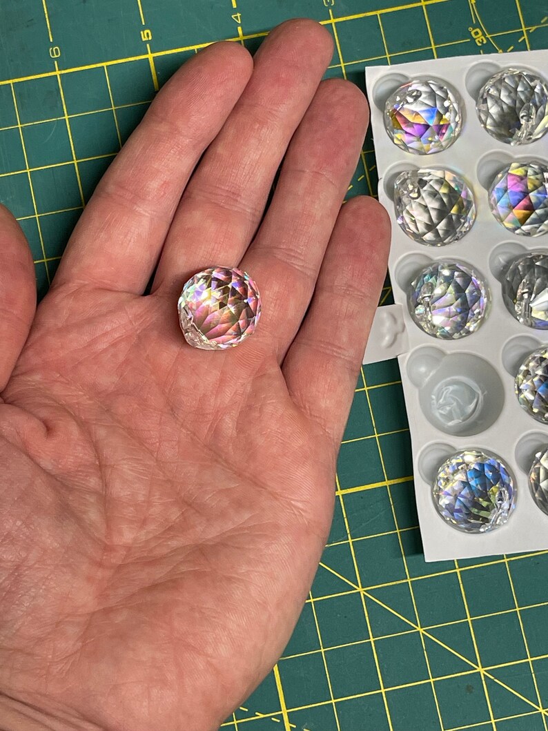 K&ouml;nnte beinhalten: Nahaufnahme einer klaren Kristallkugel mit Regenbogenfacetten. Die Kugel wird in einer Hand gehalten. Die Kugel ist Teil eines Sets von Kristallkugeln in einer wei&szlig;en Verpackung.