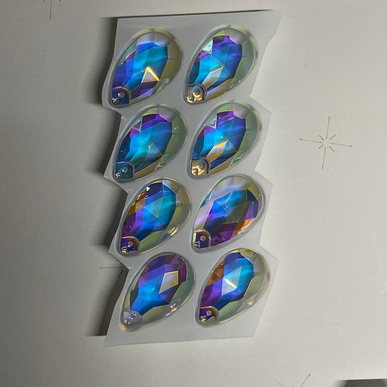 May include: A sheet of clear plastic with seven iridescent teardrop-shaped rhinestones. The rhinestones are arranged in a staggered pattern.