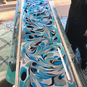 May include: A close-up of a marbled paper art piece in a wooden frame. The paper is a swirling mix of blue, black, white, and gray colors. A white tool is used to create the marbling effect.