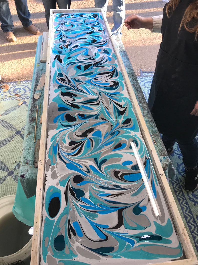 May include: A close-up of a marbled paper art piece in a wooden frame. The paper is a swirling mix of blue, black, white, and gray colors. A white tool is used to create the marbling effect.