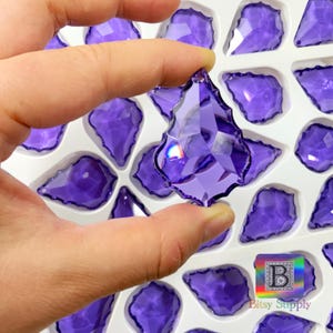 Peut inclure: Un cristal violet en forme de goutte d'eau, facett&eacute;, tenu par une main. Le cristal est entour&eacute; d'autres cristaux similaires dans un plateau blanc. Les cristaux ont de multiples facettes qui r&eacute;fl&eacute;chissent la lumi&egrave;re. Le logo Bitsy Supply est visible.